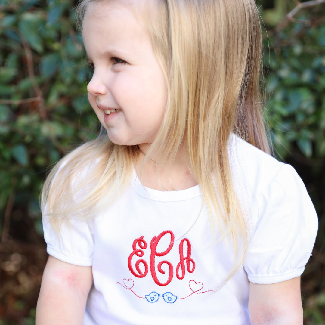 V-Day girl smiling, wearing shirt with heart embroidery.